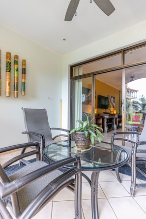 Una mesa de cristal con cuatro sillas, una planta en maceta y un ventilador de techo en una habitación con puerta corrediza de vidrio que da a un balcón.