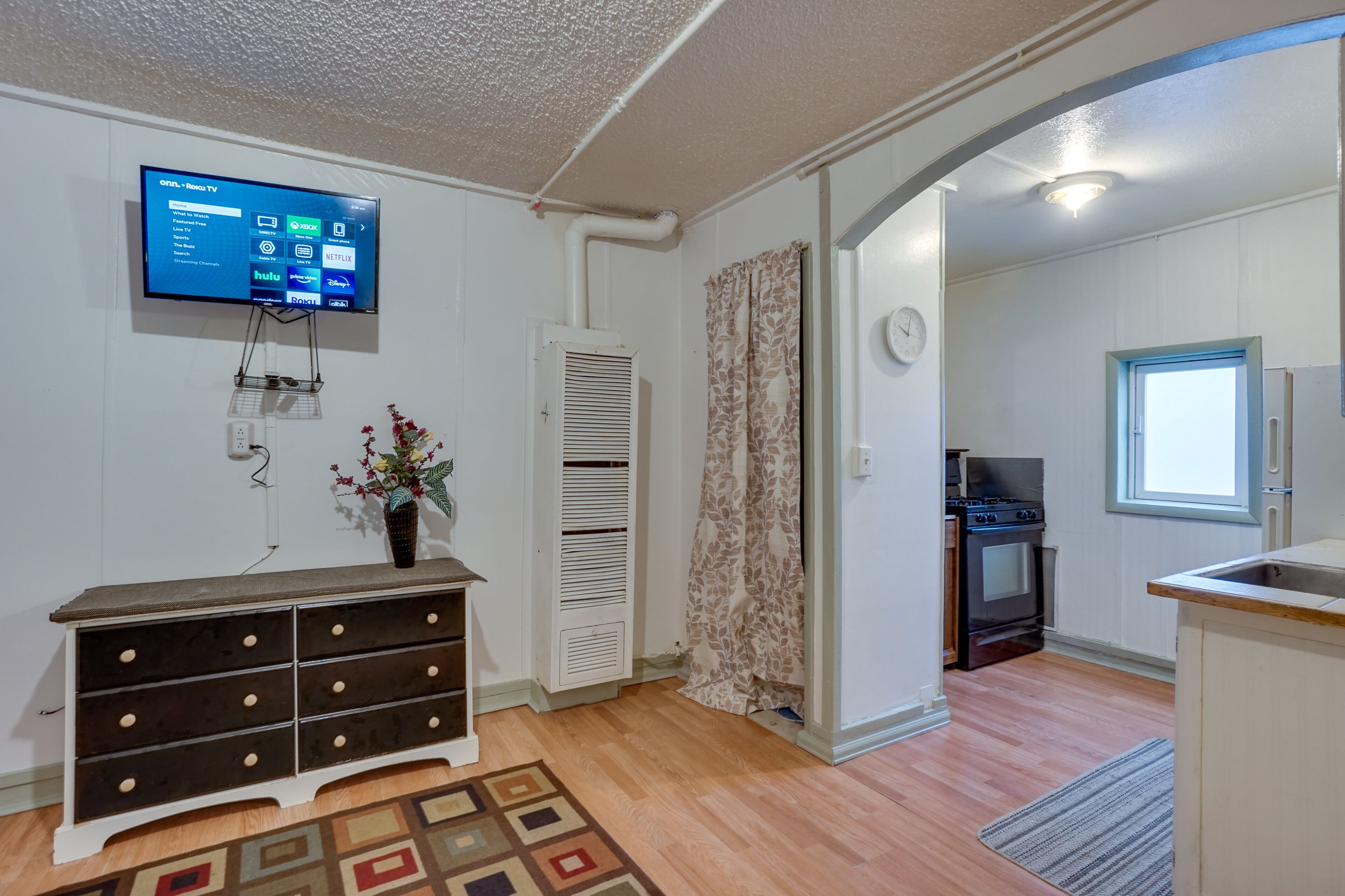 A living room with a flat-screen TV, a wooden dresser, a patterned rug, and a kitchen area with a stove and refrigerator.