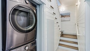 A modern laundry room with a stacked washer and dryer, a staircase with white walls, and a painting on the wall. - Sandpoint Seaclusion with PRIVATE HEATED POOL,OCEAN VIEWS,ELEVATOR& Beach Access (Port Aransas)