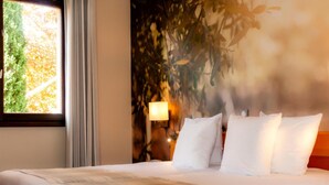Habitación de hotel con cama, ventana con cortinas, lámpara de pared y vistas a árboles.