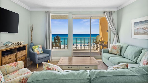 Un salon avec une grande télévision, un fauteuil bleu, un canapé vert et une table basse en bois. Il y a un balcon avec vue sur la plage et l’océan.