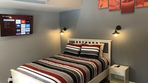 Una cama bien hecha con ropa de cama a rayas, una mesita de noche blanca y un televisor de pantalla plana montado que muestra diferentes servicios de transmisión.