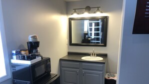 Un baño pequeño con un refrigerador negro, una cafetera, un lavamanos y un espejo.