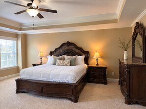 A bedroom with a large bed, two bedside tables with lamps, a ceiling fan, a dresser, and a mirror.