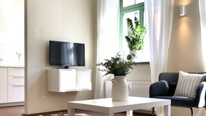 A living room with a flat-screen TV mounted on a white cabinet, a white table, a grey sofa, and a potted plant on a windowsill.