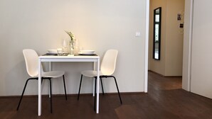 A dining area with a white table set for two, two white chairs, a vase with a plant, and a wall-mounted light fixture.