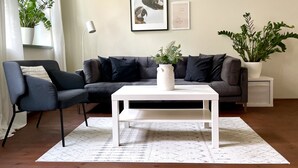 A modern living room with a dark grey sofa, a white coffee table, and a white side table. There are two potted plants, one on the sofa and one on the side table. The room has a white floor and a patterned rug.