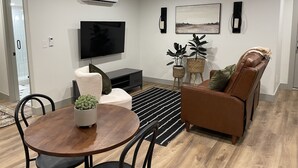Una sala de estar moderna con un televisor de pantalla plana, un sofá de cuero marrón, una mesa de comedor redonda de madera y dos plantas en macetas.