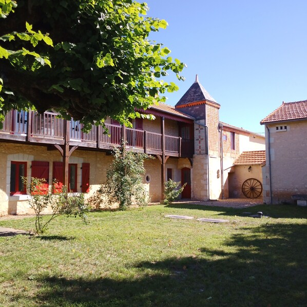 A courtyard with a building featuring a tower, wooden balconies, and a stone wall with a large wheel.
