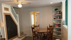 A living room with a yellow sofa, a dining table with chairs, a staircase, and a bookshelf. - Just a few blocks from Lake Erie!  4 season home with a cozy cottagey feel. (Lakeside Marblehead)