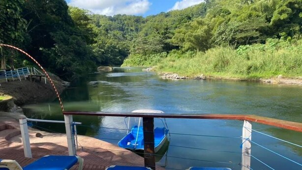 A deck with blue lounge chairs and a small blue boat, overlooking a river with a forested hillside in the background. - Tapioca Village Retreat (Castleton)
