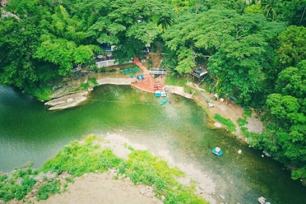 An aerial view of a riverside area with boats, a wooden bridge, and lush greenery.