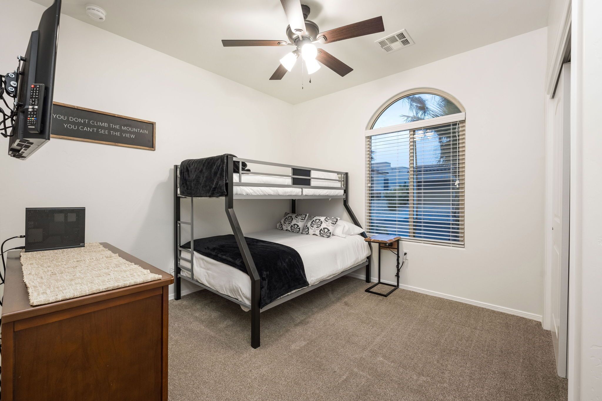 Une chambre avec un lit superposé, un bureau avec un ordinateur portable, un ventilateur de plafond et une fenêtre avec des stores.