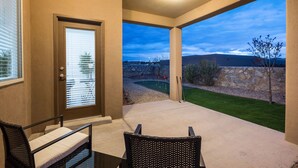 A covered patio with a view of a golf course, a door, and two wicker chairs. - House with Community Pool and Putting Green (Las Cruces)