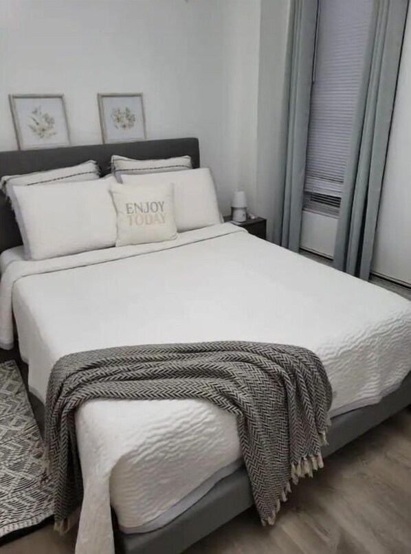Un lit bien fait, avec une couette blanche et des oreillers, un plaid à motifs chevrons gris et une table de chevet fixée au mur, sur laquelle se trouve une lampe.