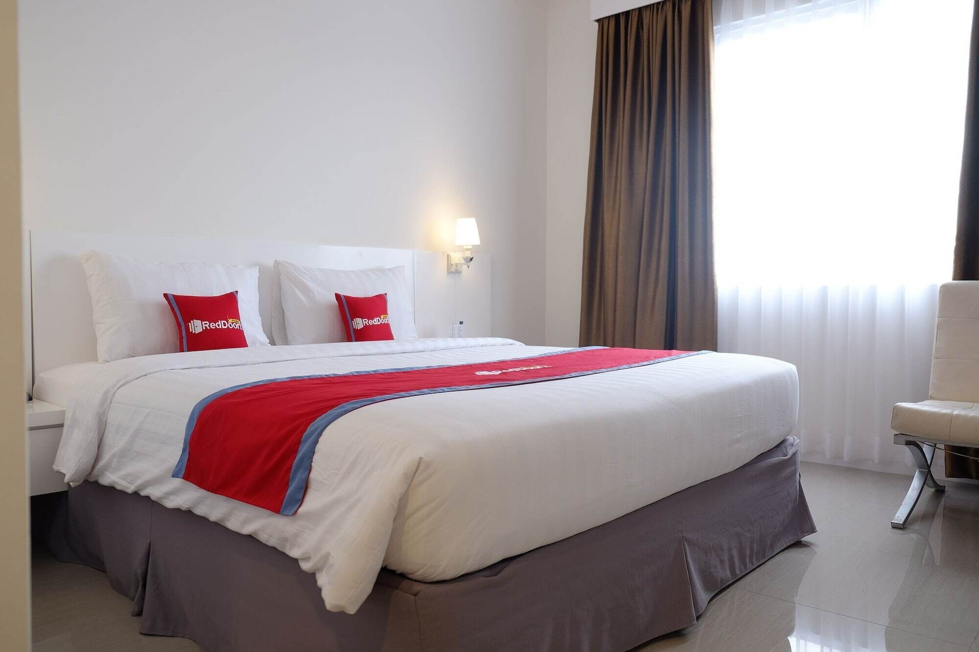 Ein Hotelzimmer mit einem großen Bett, zwei roten Kissen, einer roten und blauen Tagesdecke, einem Nachttisch, einer Lampe, einem Sessel und einem Fenster mit Vorhängen.