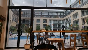 A modern indoor pool area with glass walls, wooden tables, and chairs, and a view of adjacent buildings. - Embassy Gardens by Morninglife Luxury (Accra)