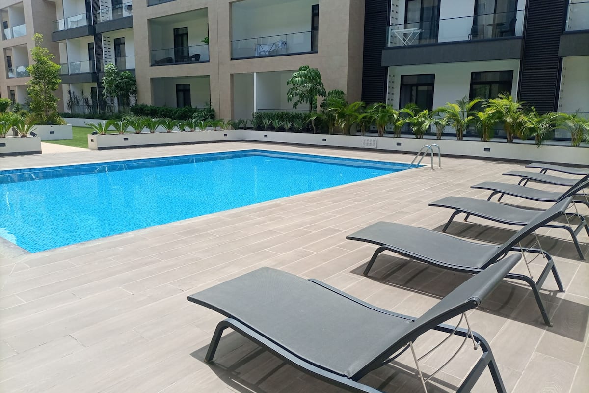 A swimming pool with lounge chairs, surrounded by a modern building with balconies and greenery.