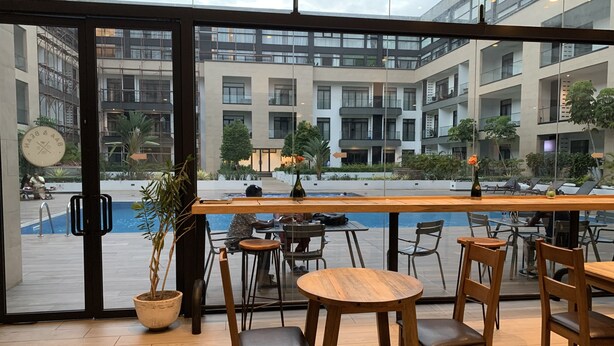 A modern outdoor seating area with wooden tables and chairs, a pool in the background, and a view of a multi-story building with balconies. - Embassy Gardens by Morninglife Luxury (Accra)