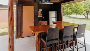 Un espace de restauration extérieur de style rustique, avec une table et des chaises en bois, une cuisine intégrée dotée d’un réfrigérateur et un téléviseur fixé au plafond.