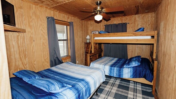 A wooden cabin room with two bunk beds, a ceiling fan, a window with curtains, and a plaid rug. - River Frontage in the Comanche Crossing Subdivision (Concan)