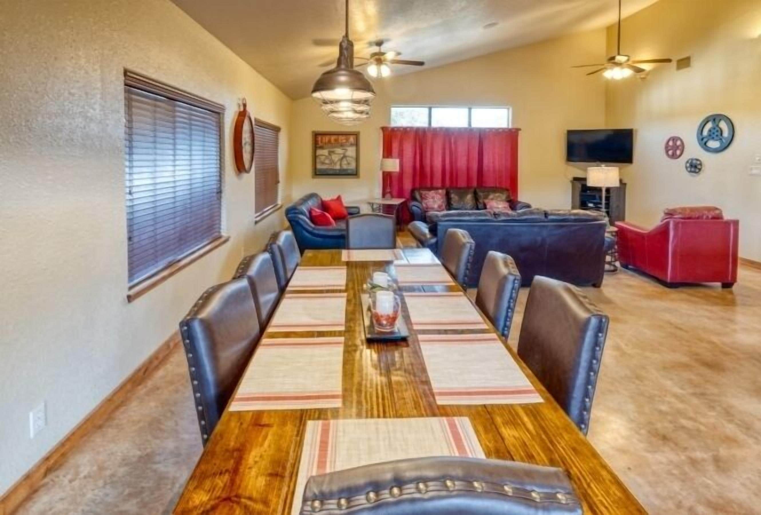 A spacious living room with a large wooden dining table, leather chairs, and a sofa. There are ceiling fans, a TV, and a red curtain.