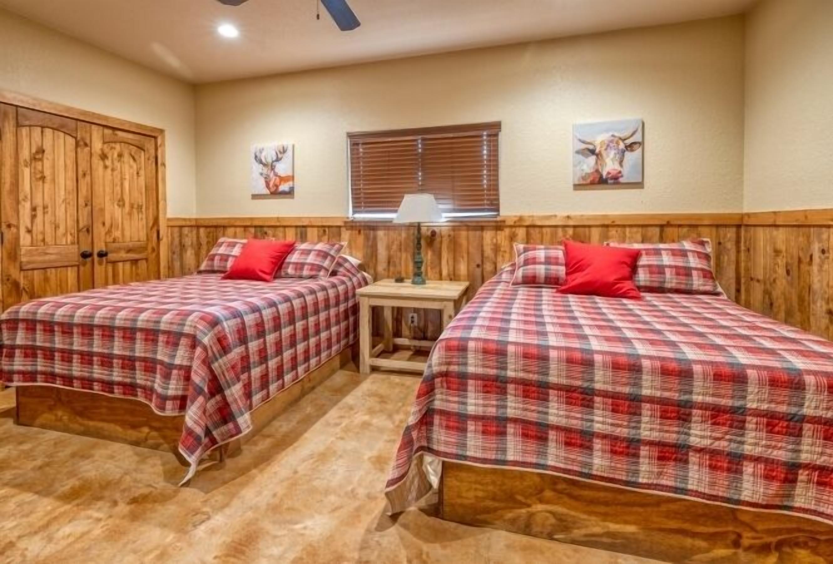A room with two beds, each with a red and white checkered bedspread and matching pillows. There is a wooden wardrobe on the left, a window with blinds, and a wall-mounted lamp.
