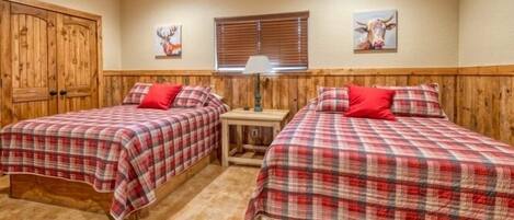 A room with two beds, each with a red and white checkered bedspread and matching pillows. There is a wooden wardrobe on the left, a window with blinds, and a wall-mounted lamp.