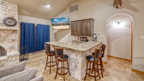 A kitchen with a stone island, bar stools, a mounted deer head, a stone fireplace, and a television above the island. - Beautiful and spacious home with a pool (Concan)