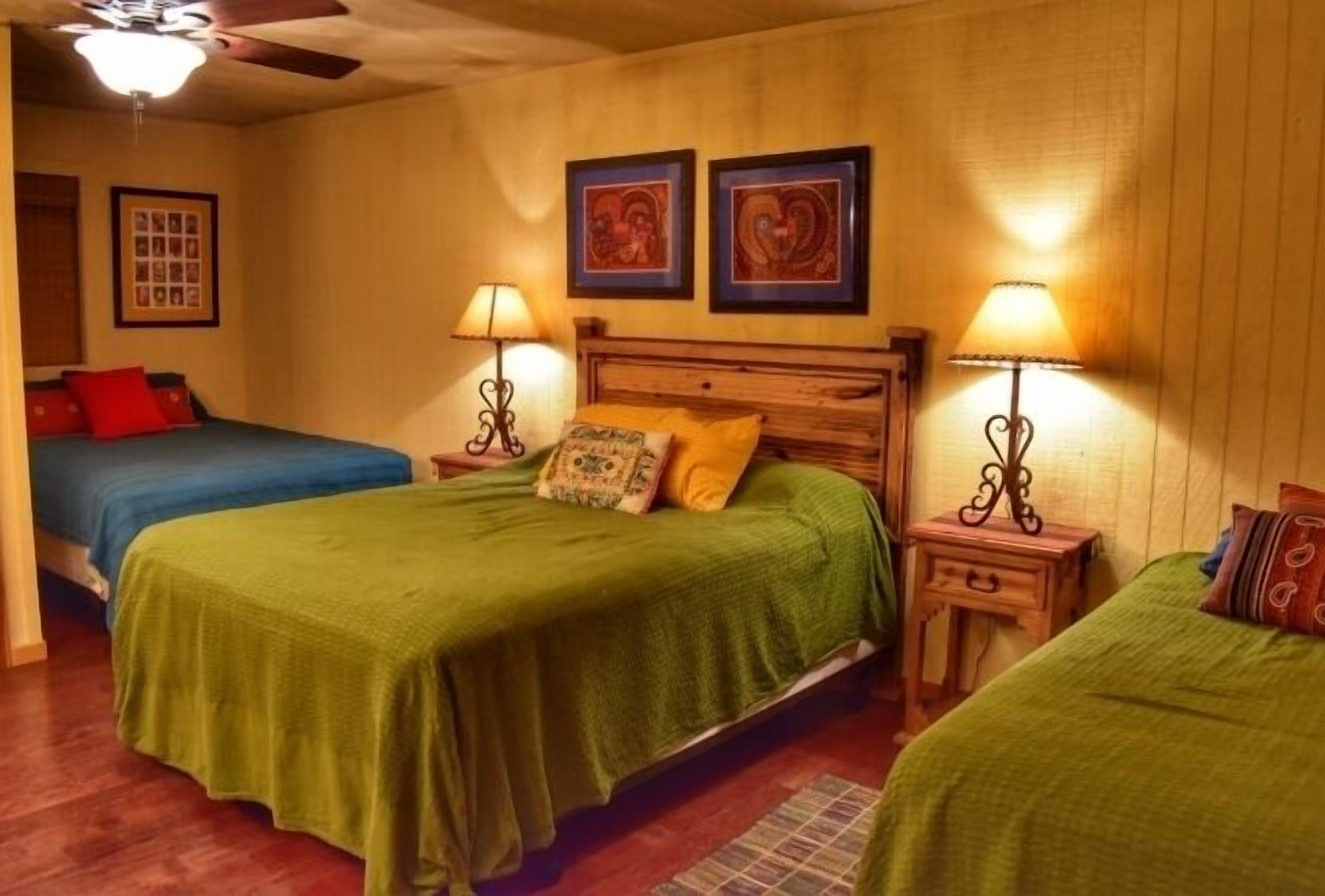 A room with two beds, each with a green bedspread and a wooden headboard. There are two bedside tables with lamps, and the walls are adorned with framed artwork.