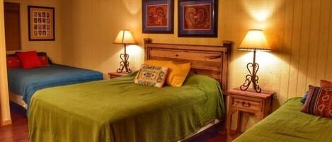 A room with two beds, each with a green bedspread and a wooden headboard. There are two bedside tables with lamps, and the walls are adorned with framed artwork.