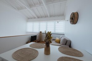 A modern, minimalist living space with a white interior, a sofa with cushions, a coffee table with woven placemats, and a vase with flowers.