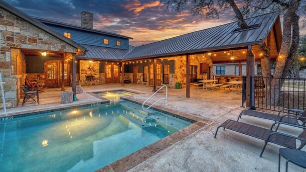 Un área de piscina con un edificio de piedra, asientos exteriores y un cielo al atardecer.