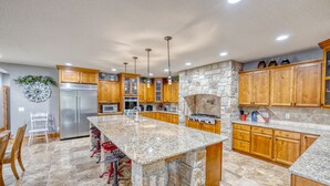 Una cocina amplia con encimeras de granito, armarios de madera, chimenea de piedra y refrigerador de acero inoxidable.