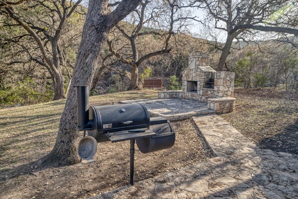 A stone fireplace and a black grill in a wooded outdoor area.