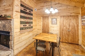 A wooden cabin interior with a stone fireplace, a large wooden table with chairs, and a hanging light fixture.