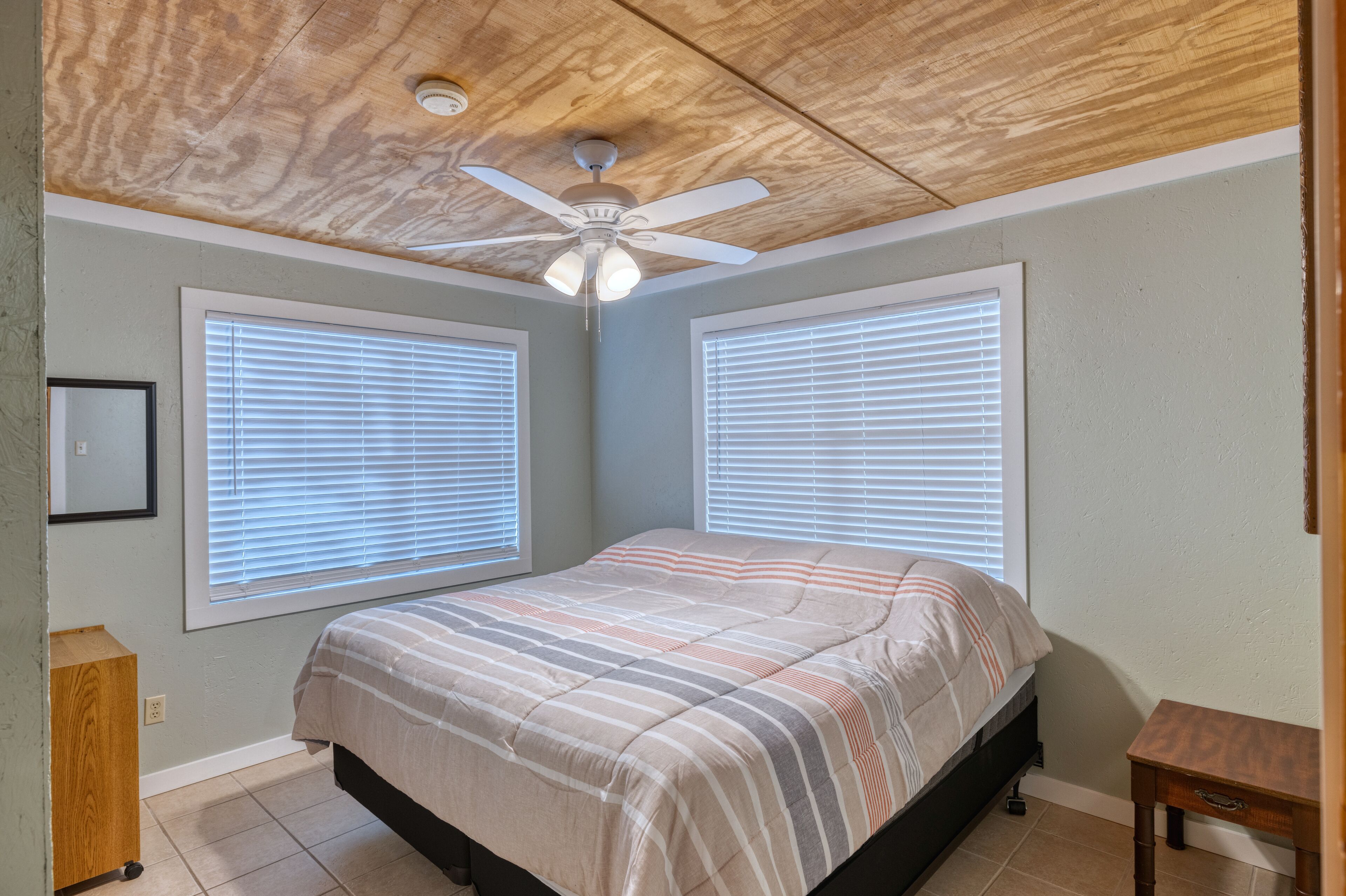 A bedroom with a bed, two windows with blinds, a ceiling fan, and a small wooden nightstand.