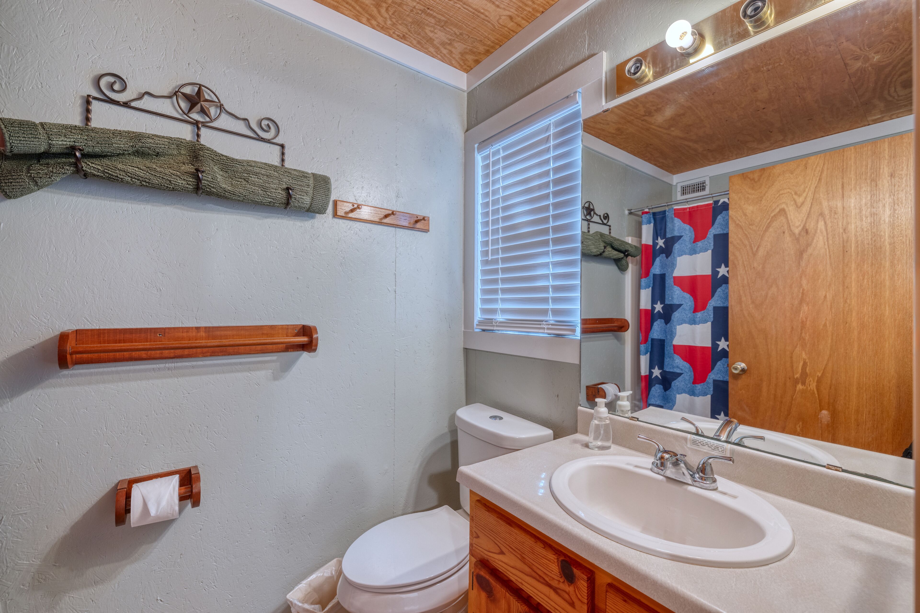 A bathroom with a toilet, sink, and mirror. There is a towel rack, a window with blinds, and a shower curtain with a flag design.