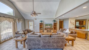 A spacious living room with a large grey sofa, wooden coffee tables, and a ceiling fan. There are wooden shelves with decorative items and a television. - Large river front home in Cypress Bend (Concan)