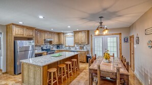 A kitchen with wooden cabinets, a stainless steel refrigerator, a granite countertop, and a dining area with a wooden table and bar stools. - Large river front home in Cypress Bend (Concan)