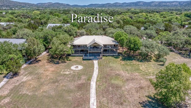 An aerial view of a large, single-story house with a prominent porch and a circular driveway, surrounded by dense vegetation and hills in the background. - Large river front home in Cypress Bend (Concan)