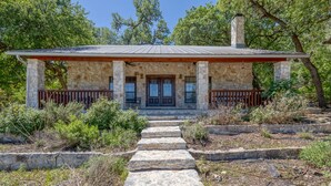 A stone building with a wooden porch, stone steps leading to the entrance, and a natural surrounding with trees and shrubs. - Beautiful home in the Comanche Crossing Subdivision (Concan)