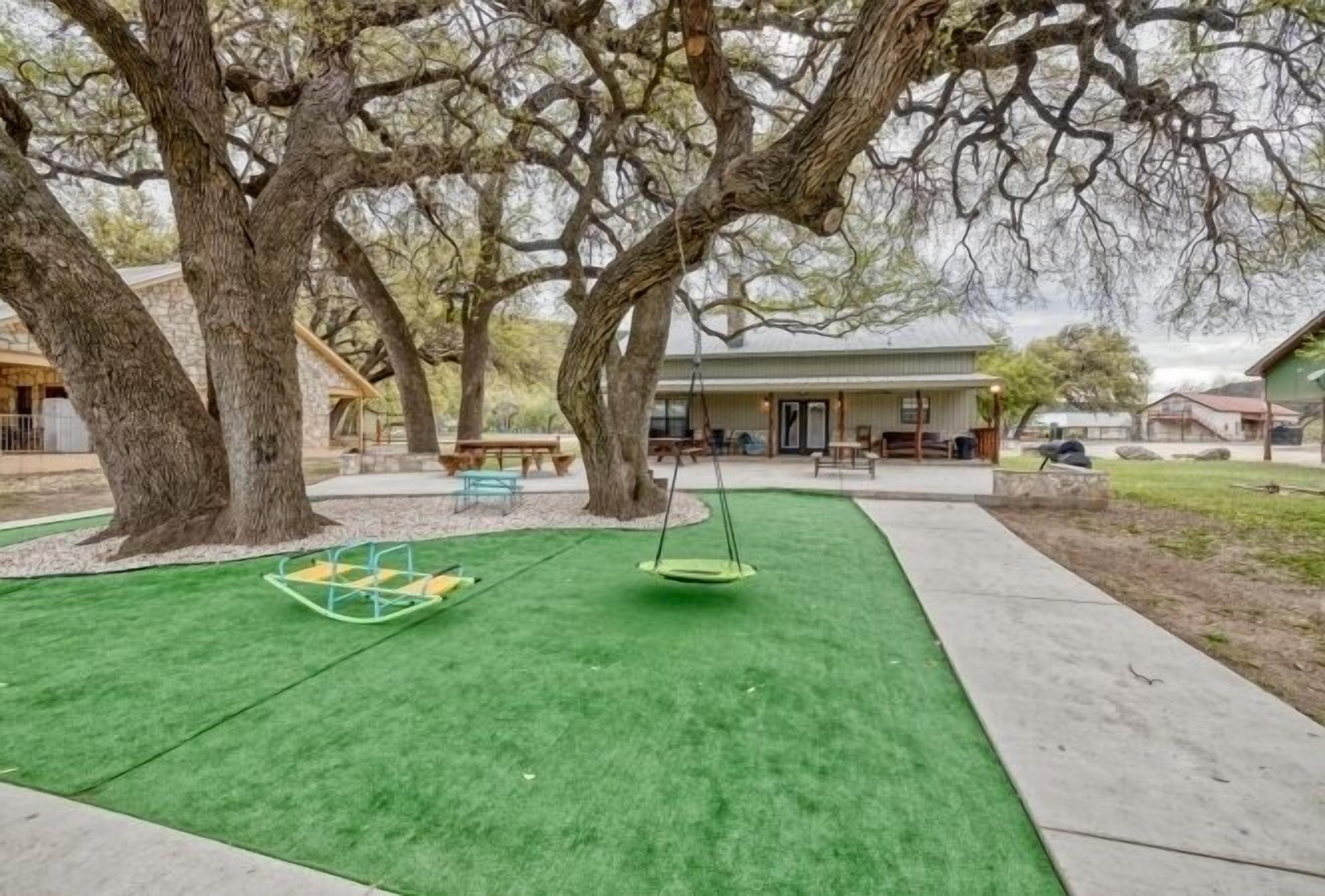A playground with a swing set and artificial turf, surrounded by trees and a building in the background.