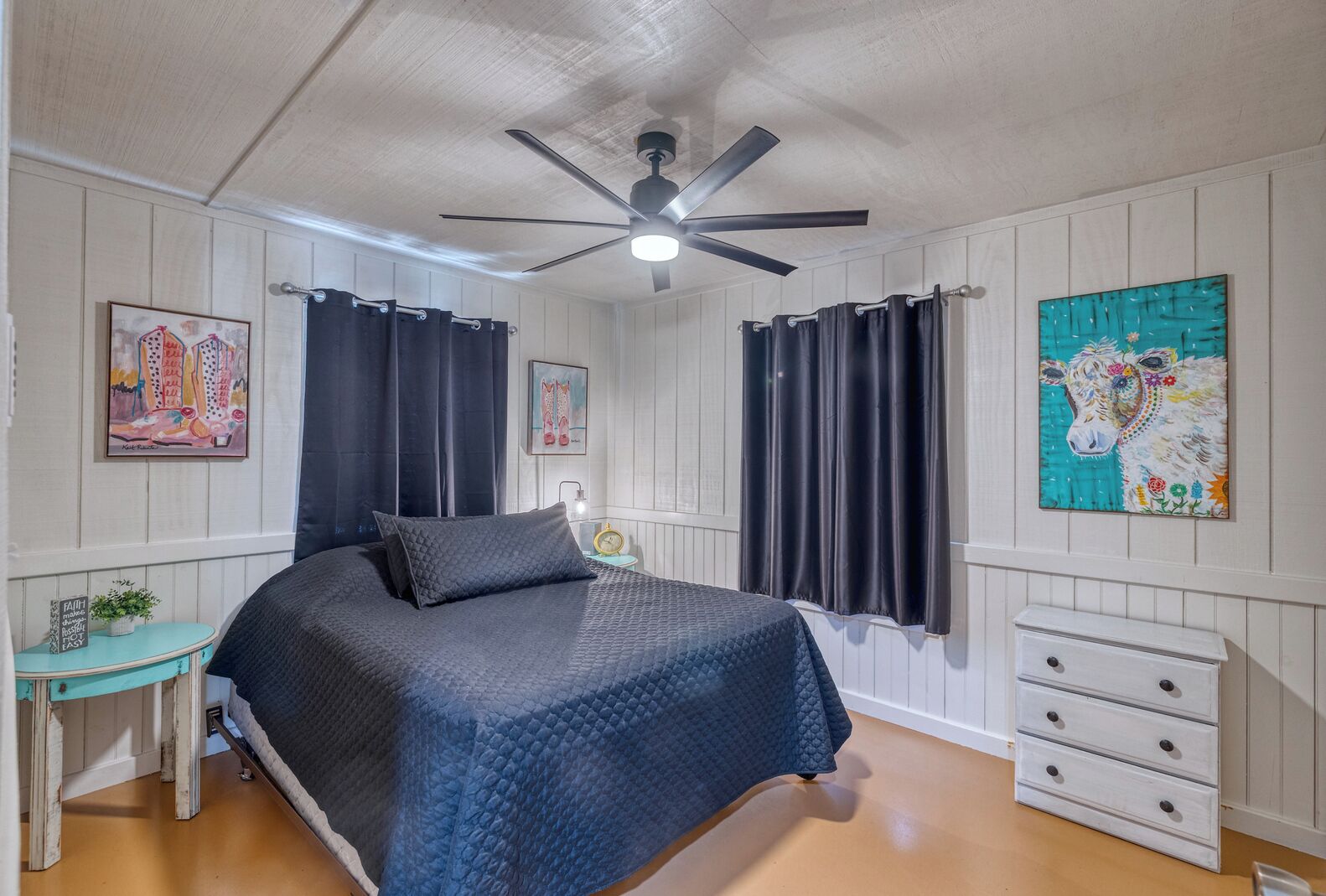 A bedroom with a bed, a ceiling fan, a nightstand, a dresser, and artwork on the walls.