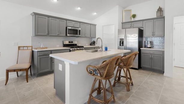 A modern kitchen with grey cabinets, stainless steel appliances, a central island with bar stools, and a dining area with a chair.