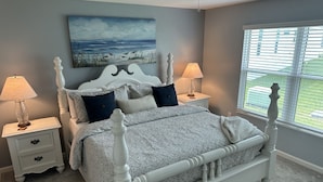 A bedroom with a white four-poster bed, two bedside tables with lamps, a painting on the wall, and a window with blinds.