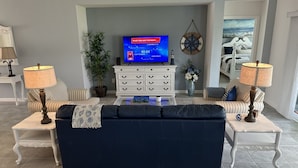 A living room with a dark blue sofa, two side tables with lamps, a white entertainment unit with a TV displaying a show, a potted plant, and a wall-mounted compass.