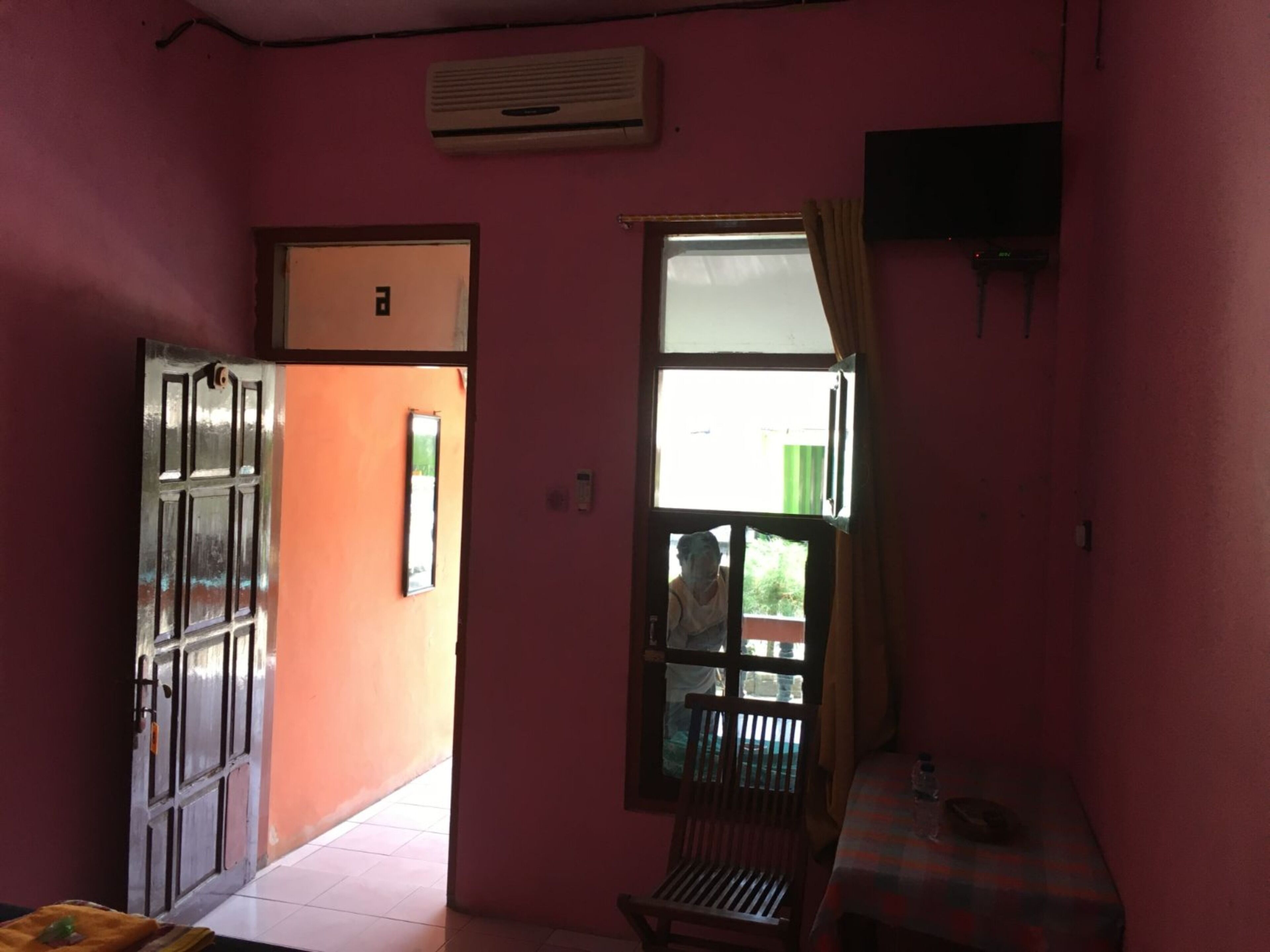 Una habitación con paredes rosas, una ventana con cortinas, una mesa con una botella y una puerta que da al exterior.