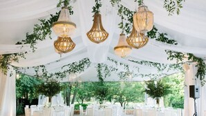 Un lieu de réception en plein air pour les mariages, avec des tables et des chaises blanches, des lustres suspendus et de la verdure.