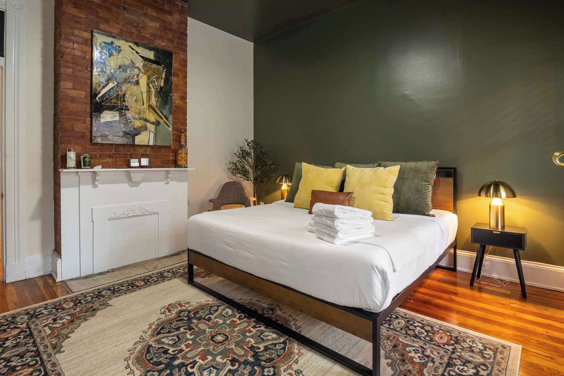 A bedroom with a bed, a nightstand, a lamp, a wall-mounted shelf with items, and a patterned rug.